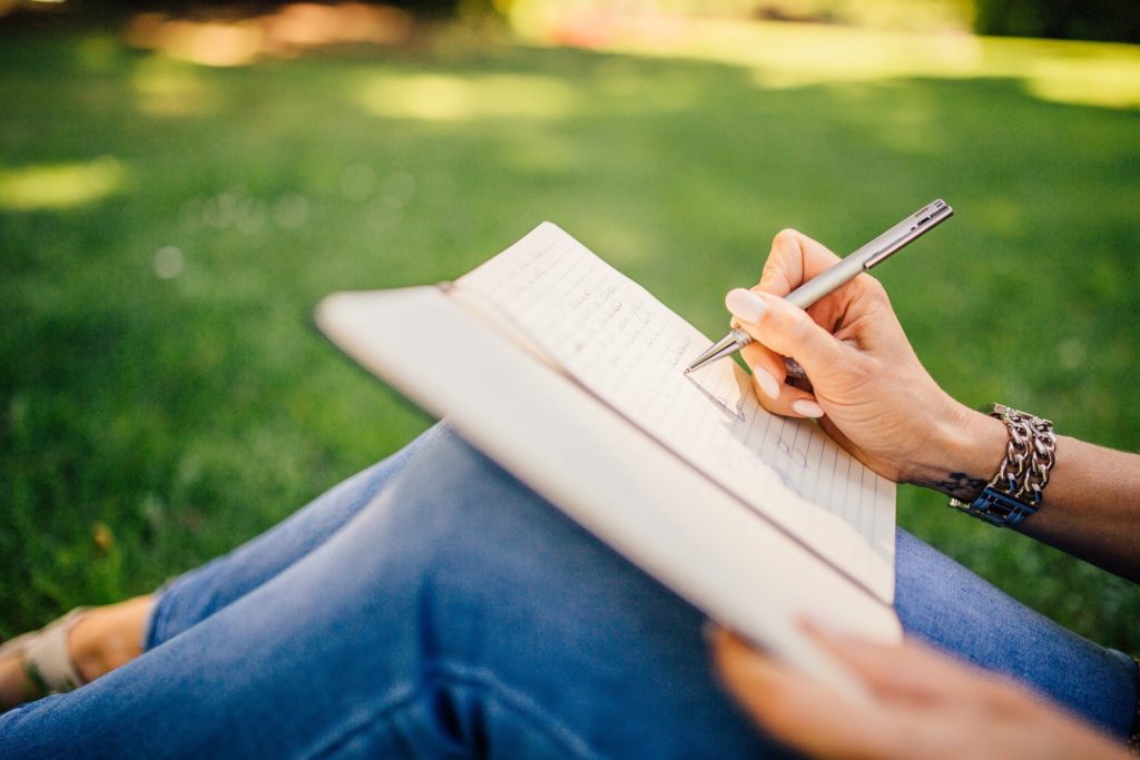 picture of woman in jeans, sitting on the grass outside, writing in a notebook; writing is therapy.